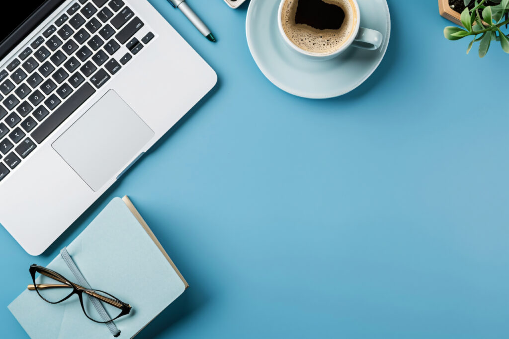 Imagen decorativa de vista desde arriba de una mesa de oficina azul con un portatil, una taza de cafe, una libreta y unas gafas de vista